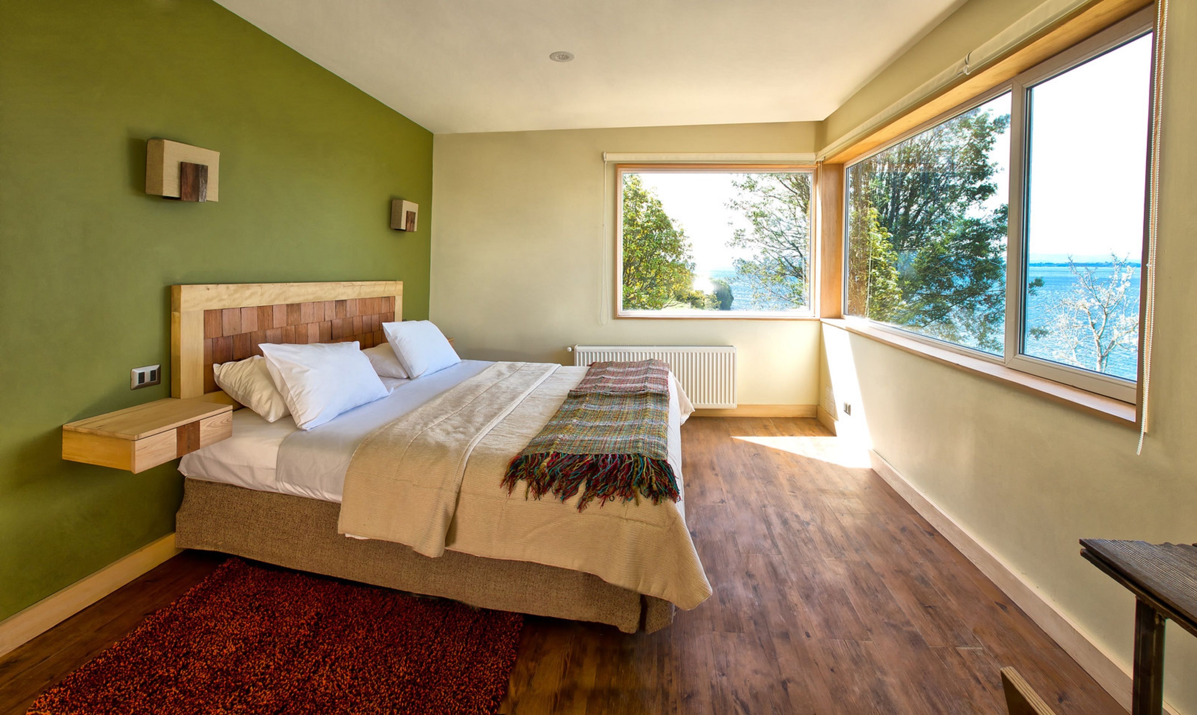 Quarto com cama de madeira, tapete vermelho, janela com vista para árvores e radiador branco.