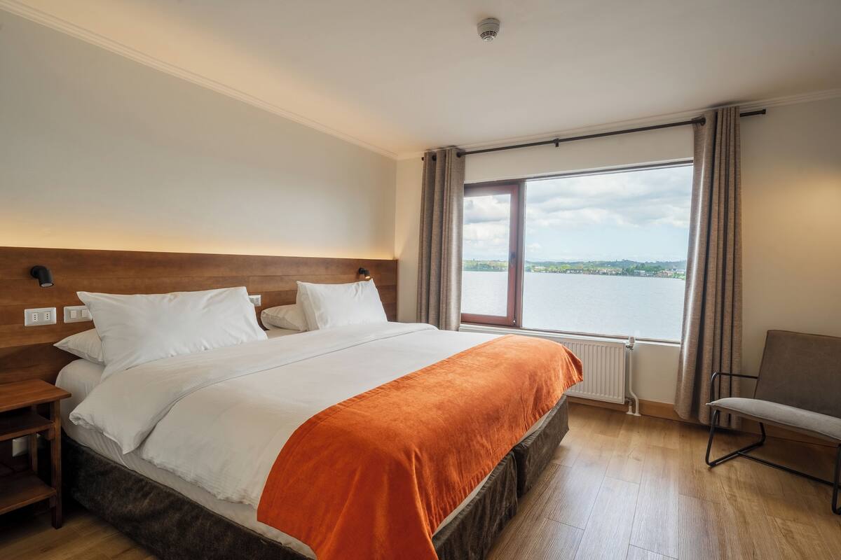 A hotel room with a large bed, a view of a lake, and a chair by the window.