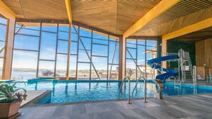 Piscina interna com vista para uma ponte e edifícios, teto de madeira e um escorrega azul.