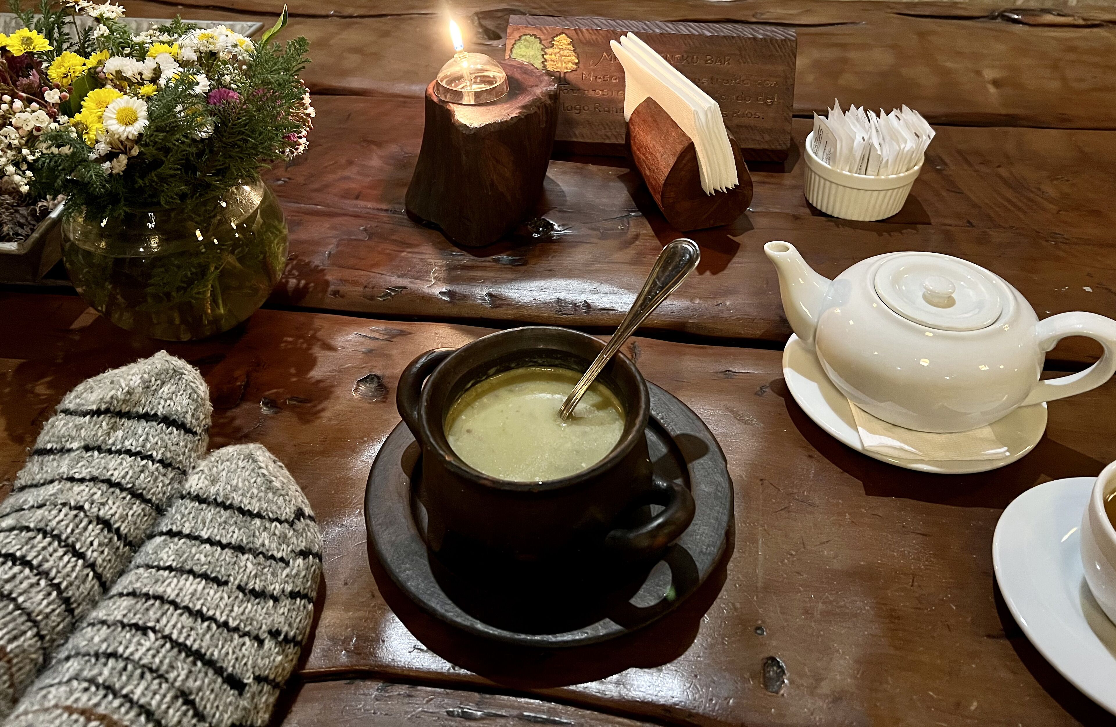 Una mesa de madera con un cuenco de sopa, una tetera, una vela, un florero con flores y un par de guantes de punto.