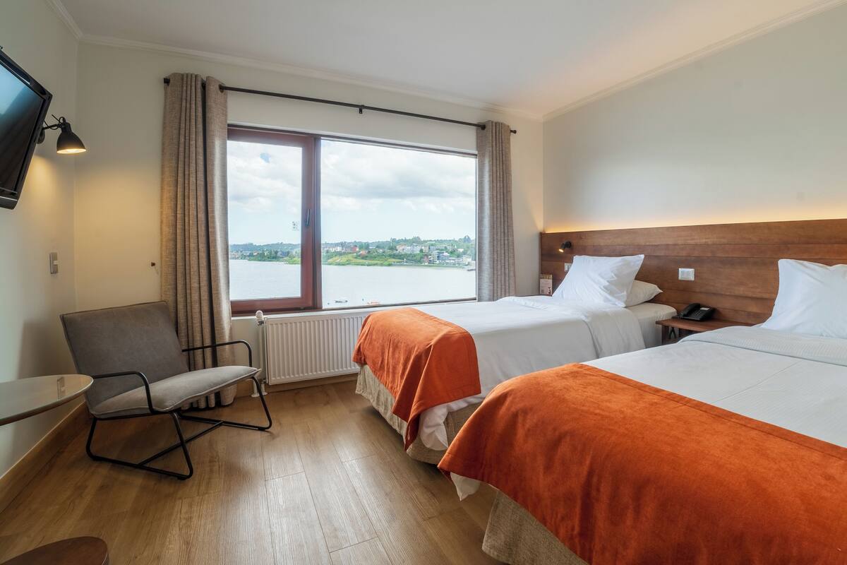 A hotel room with two beds, a chair, a TV, and a window with a view of a body of water and buildings.