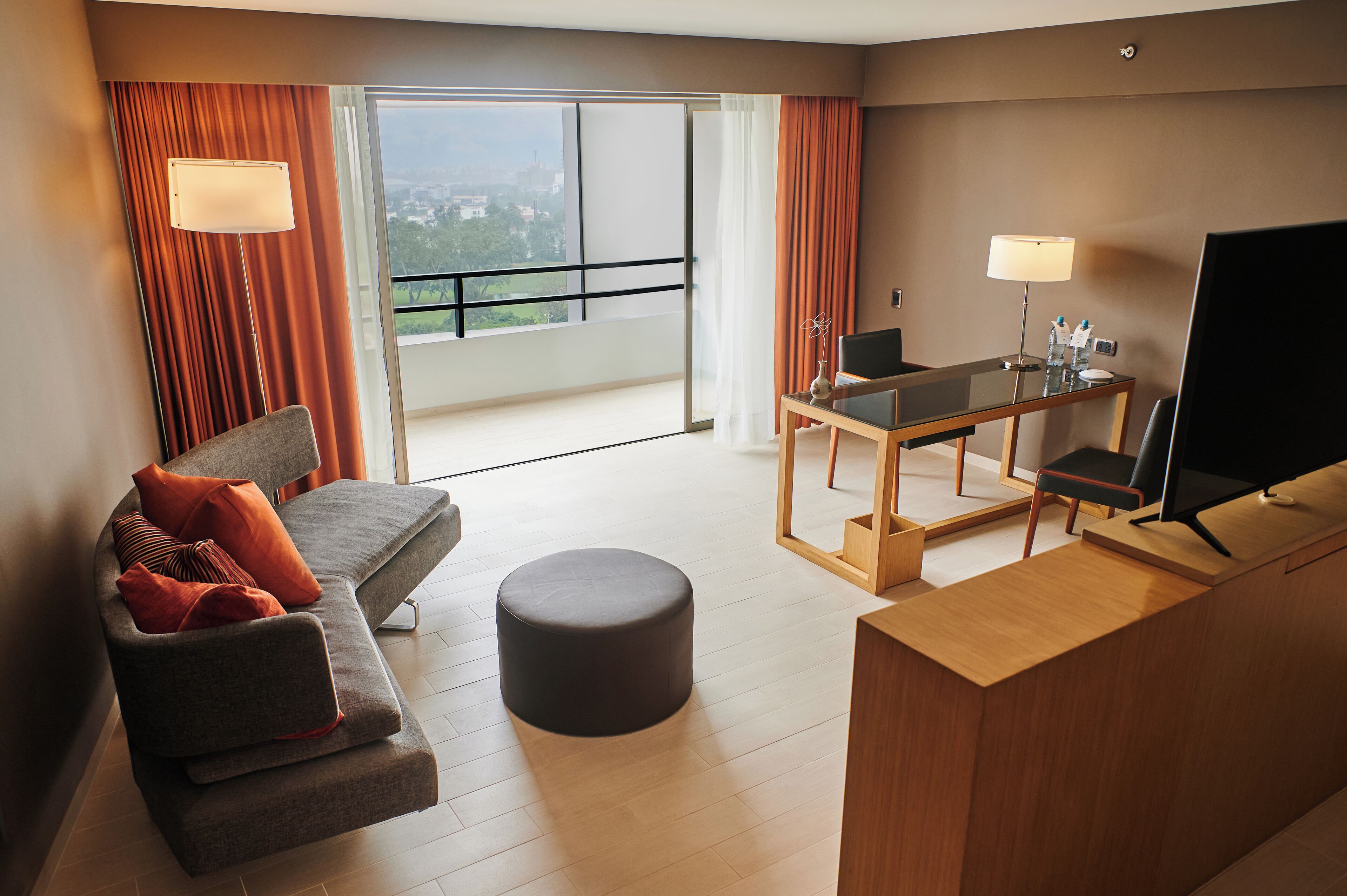 Habitació d'hotel moderna amb sofà, otomana, escriptori, cadira, televisió i un balcó amb cortines.