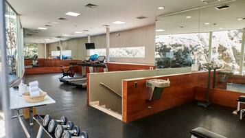 Un gimnasio con equipo de cardio, bancos de pesas y un amplio ventanal con vista a árboles.