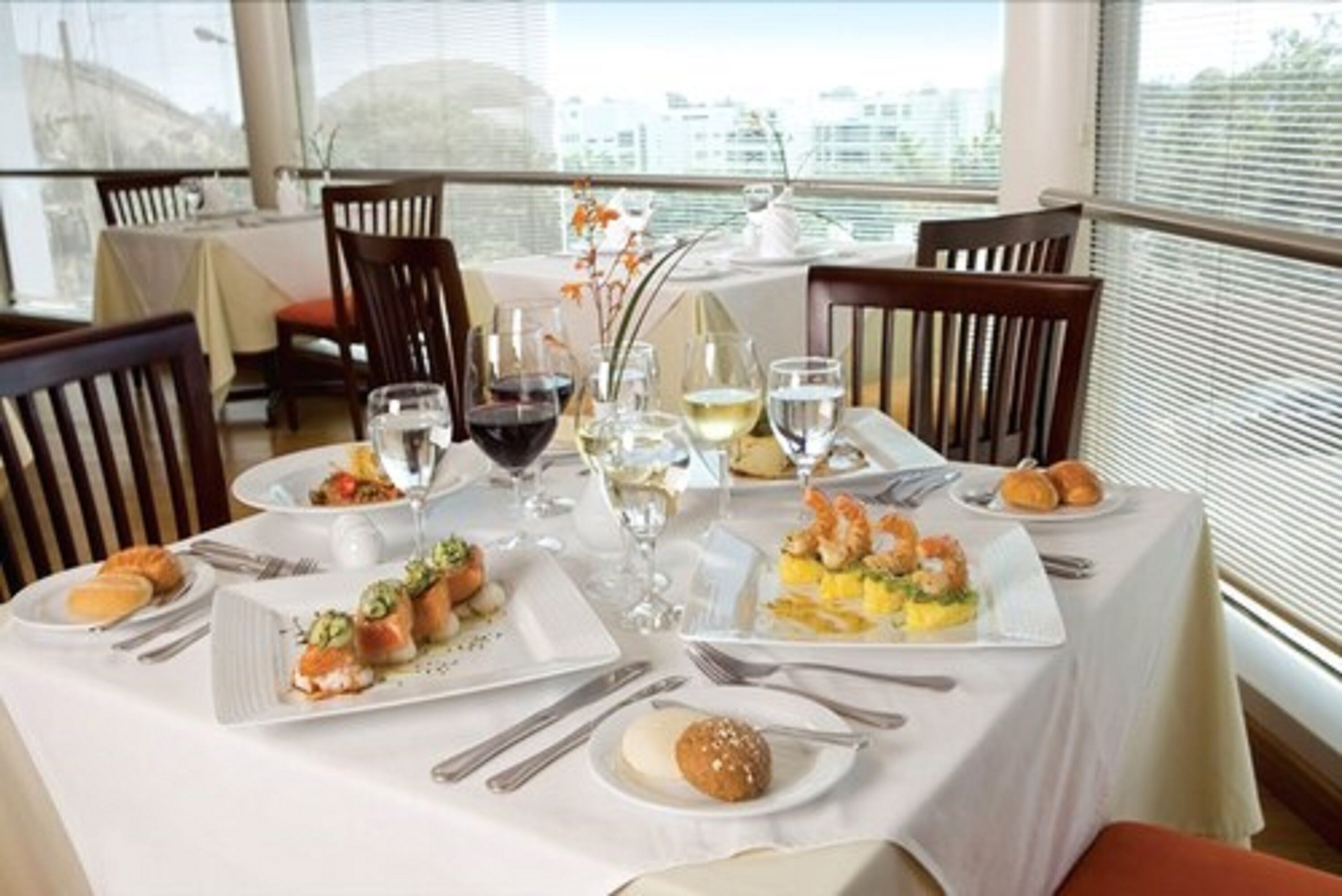 Una mesa de comedor dispuesta con diversos platos y bebidas, rodeada de sillas, en una habitación con ventanas amplias y vistas a edificios.