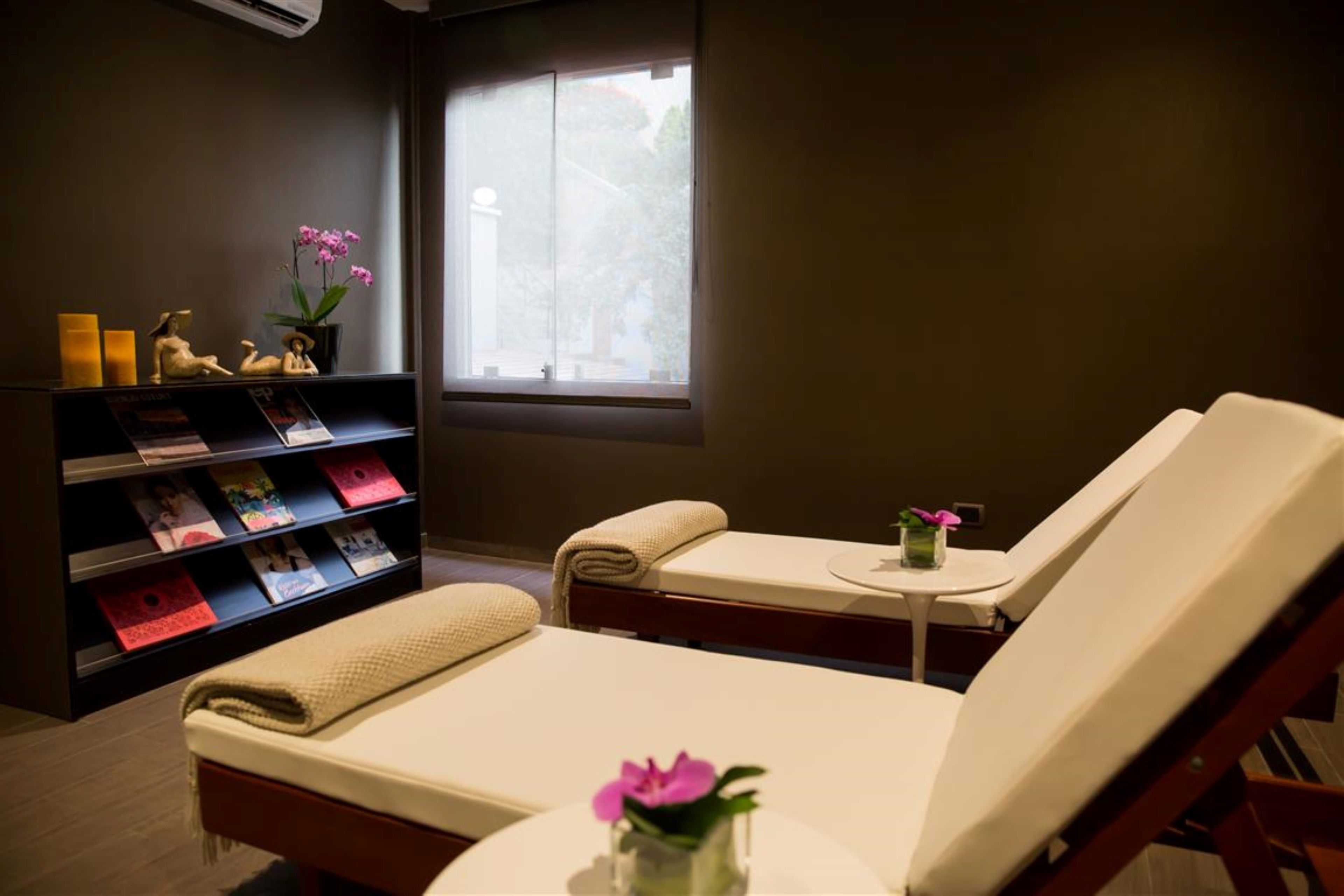 A spa room with a bed, a small table with a vase of flowers, a bookshelf with magazines, and a window with a view.