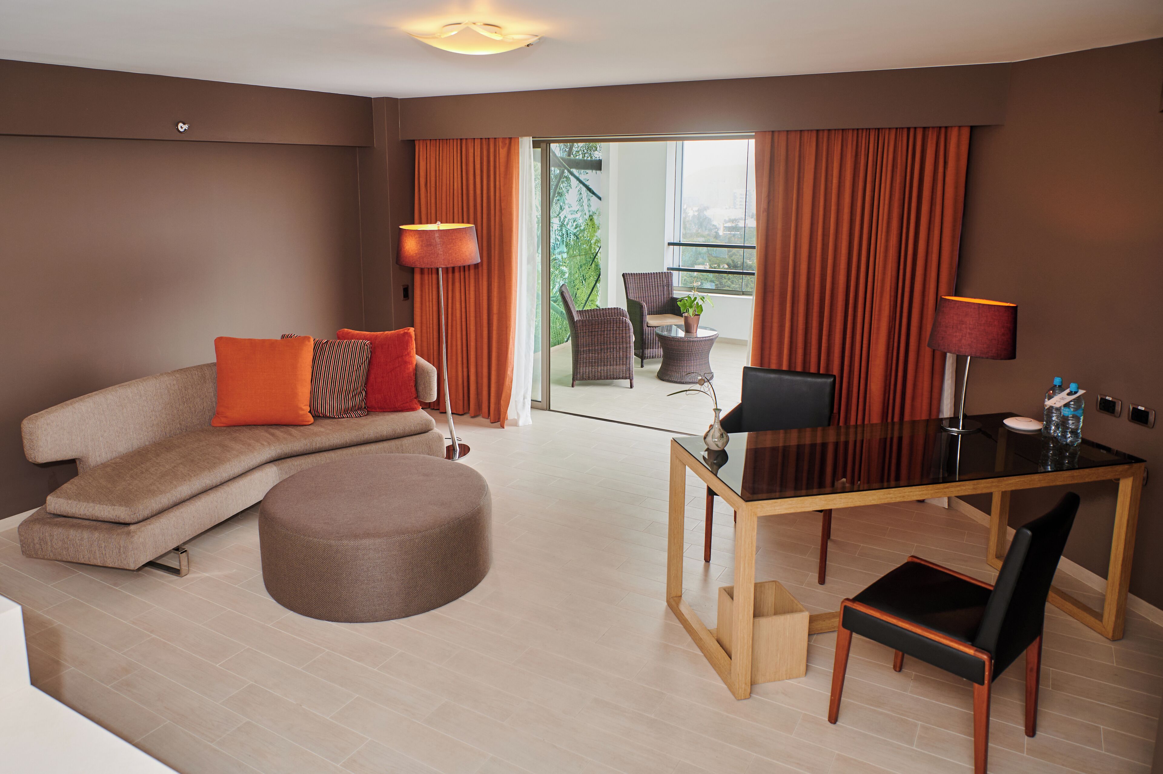 A modern living room with a sofa, ottoman, dining table, and chairs. There is a balcony with curtains and a view of greenery.