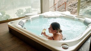 A woman in a hot tub with a glass of white wine, wooden flooring, and a view of a rocky landscape through large windows. - Casa Andina Premium Golf Los Incas (Santiago de Surco)