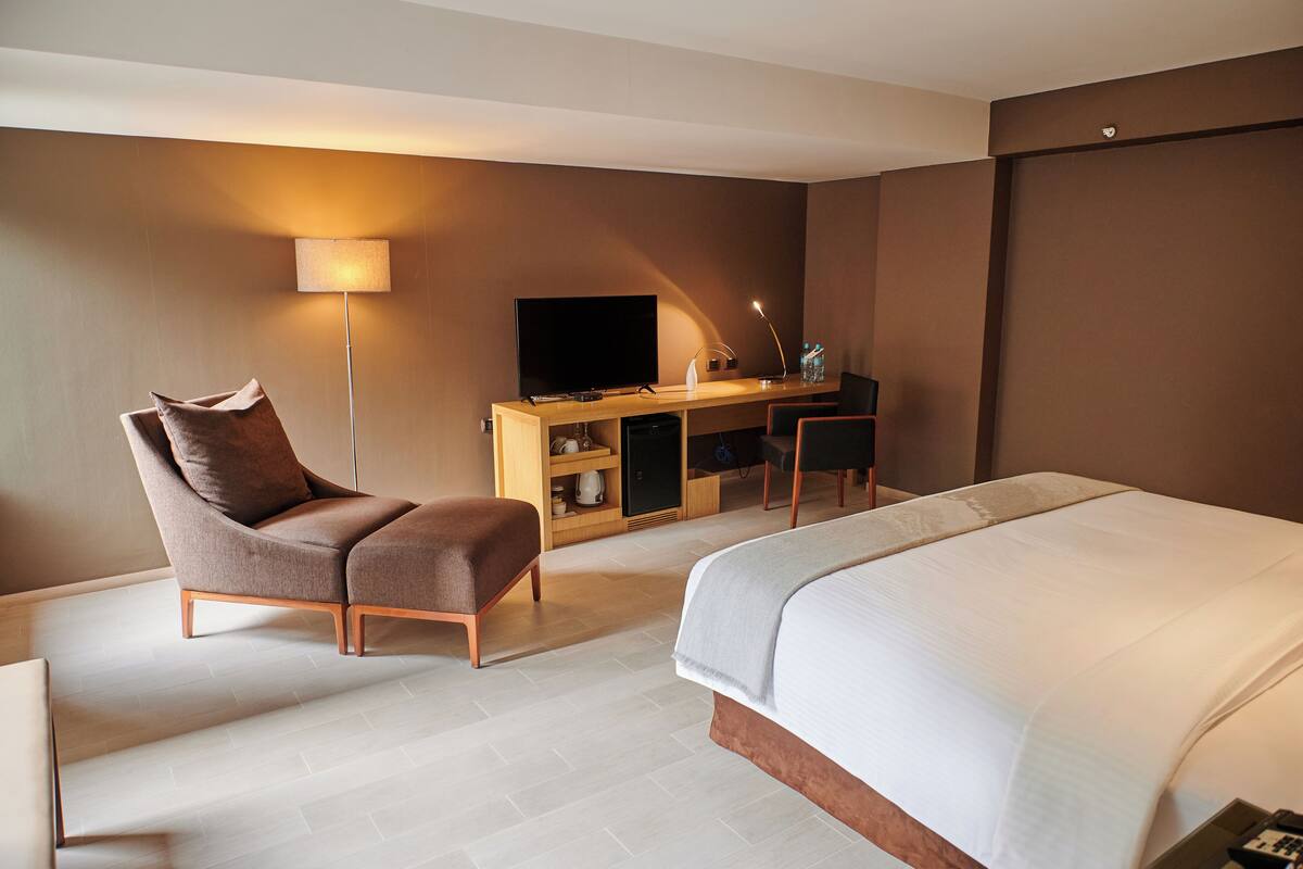 A modern hotel room with a large bed, a desk with a flat-screen TV, a chair, and a lamp.