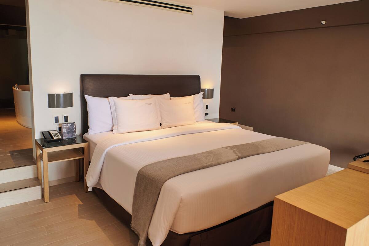 A neatly made bed with white linens and a dark headboard, two bedside lamps, and a wooden nightstand with a phone and a box.