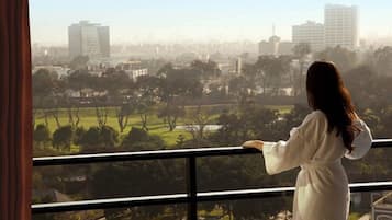 Una persona vestida de blanco de pie junto a una ventana con vistas a un paisaje urbano y zonas verdes.