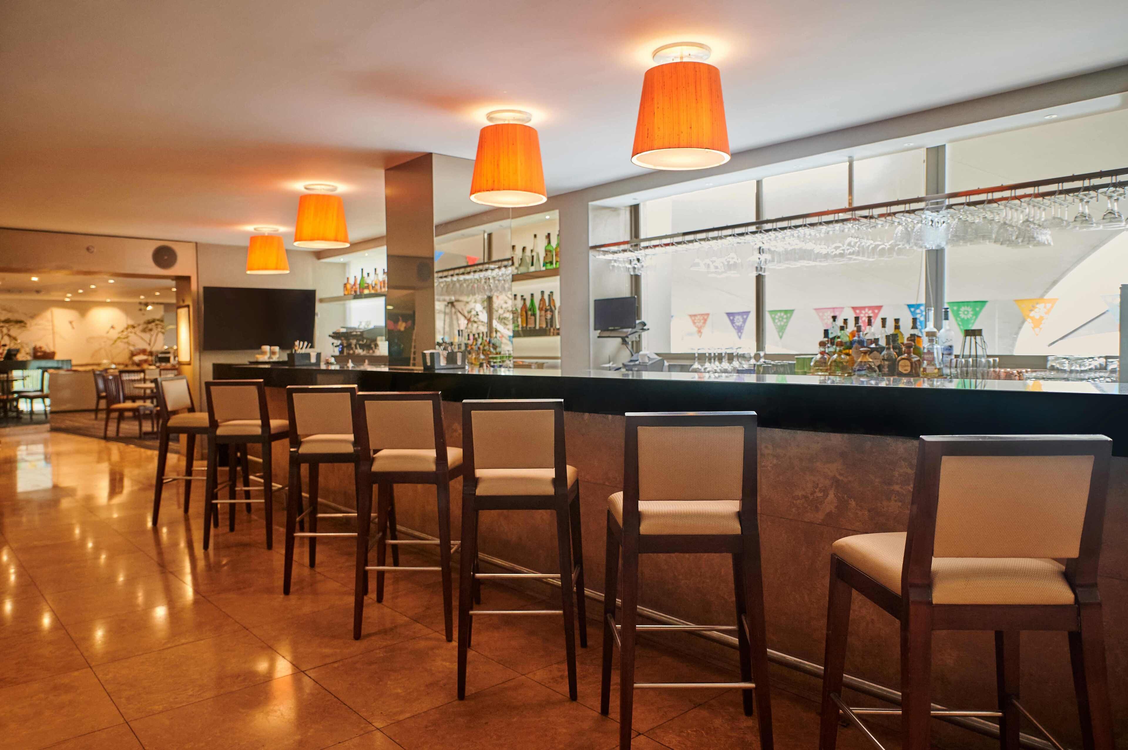 Zona de bar con sillas de madera, mostrador con vitrina de vidrio y lámparas colgantes naranjas.