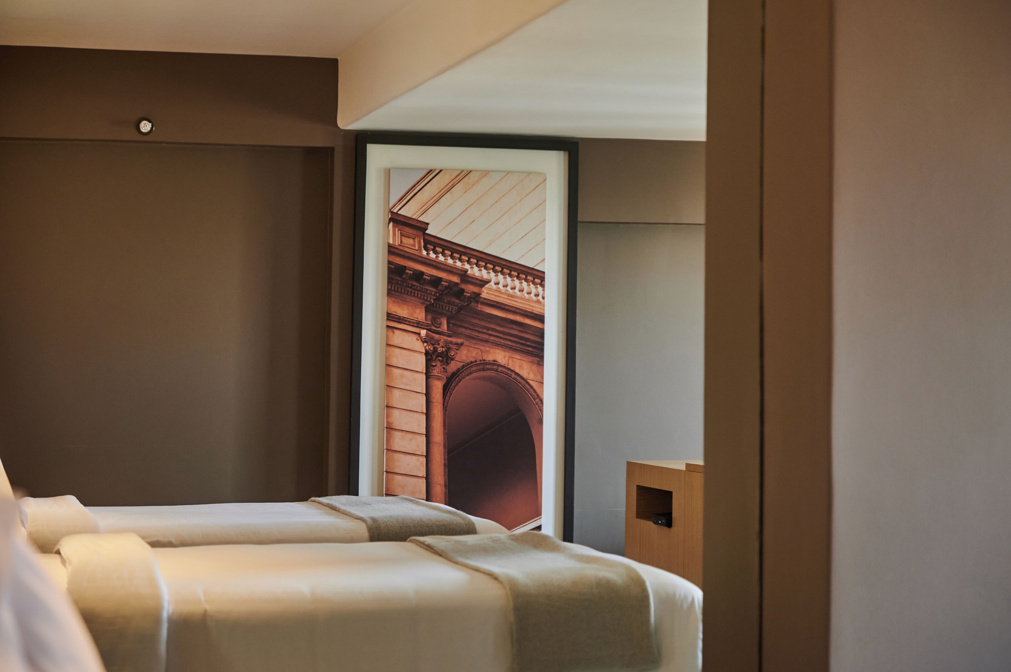 Habitación de hotel con dos camas, una mesita de noche de madera y un cuadro grande con un arco arquitectónico.
