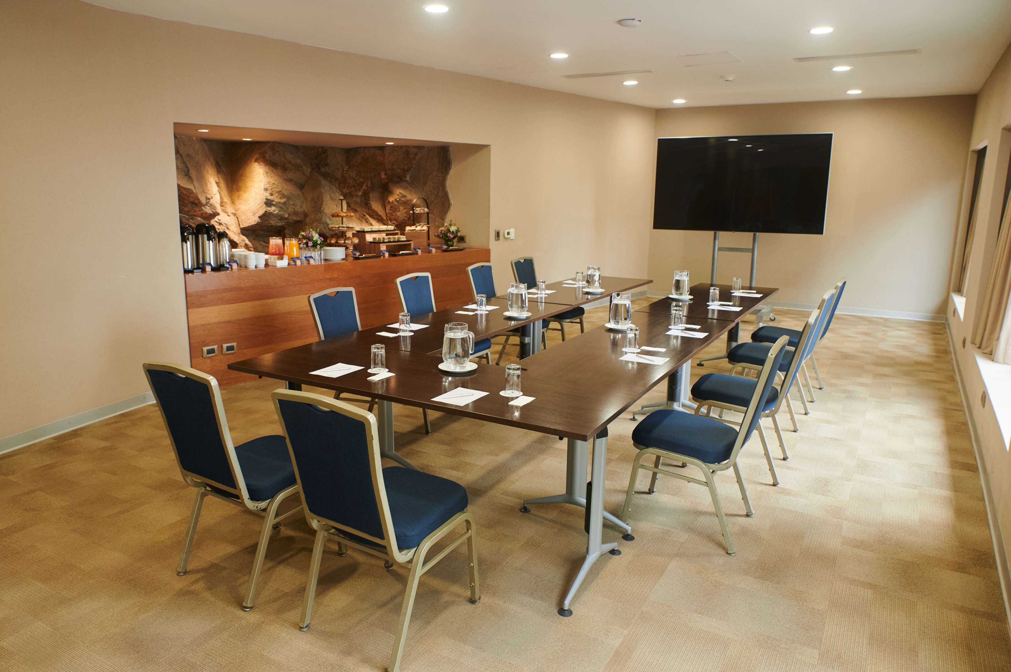 Un salón de conferencias con una larga mesa, sillas azules, una estación de café y una pantalla grande.