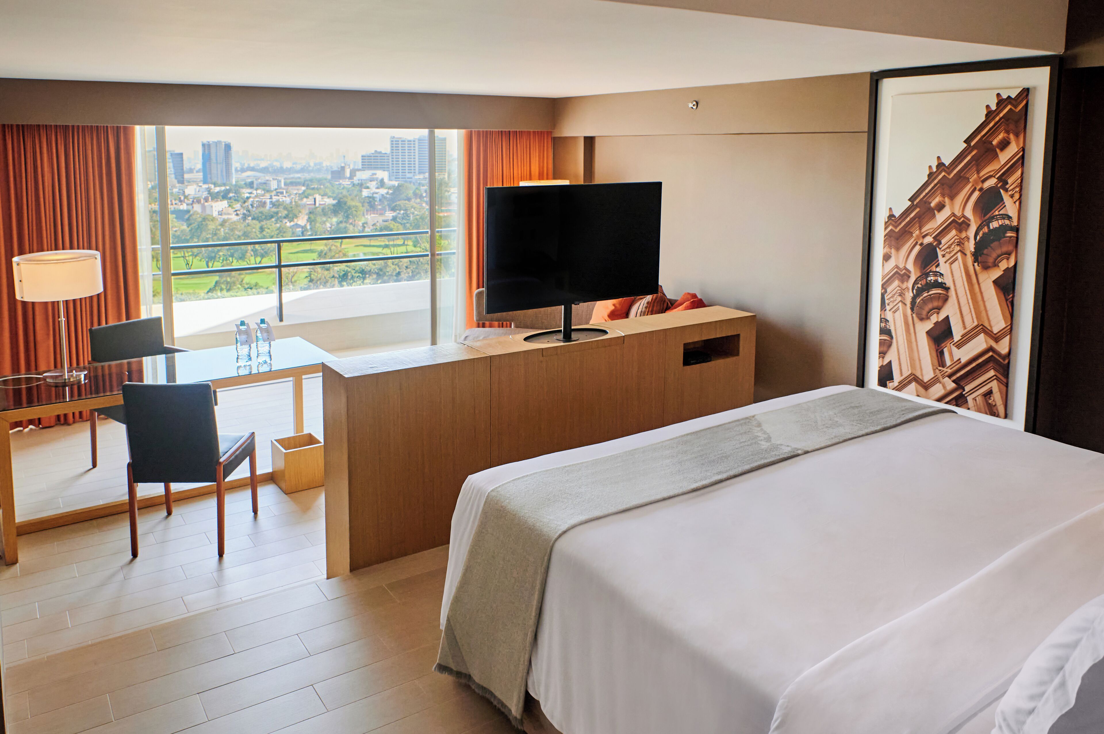 A hotel room with a bed, a desk with a TV, a chair, and a view of a cityscape.