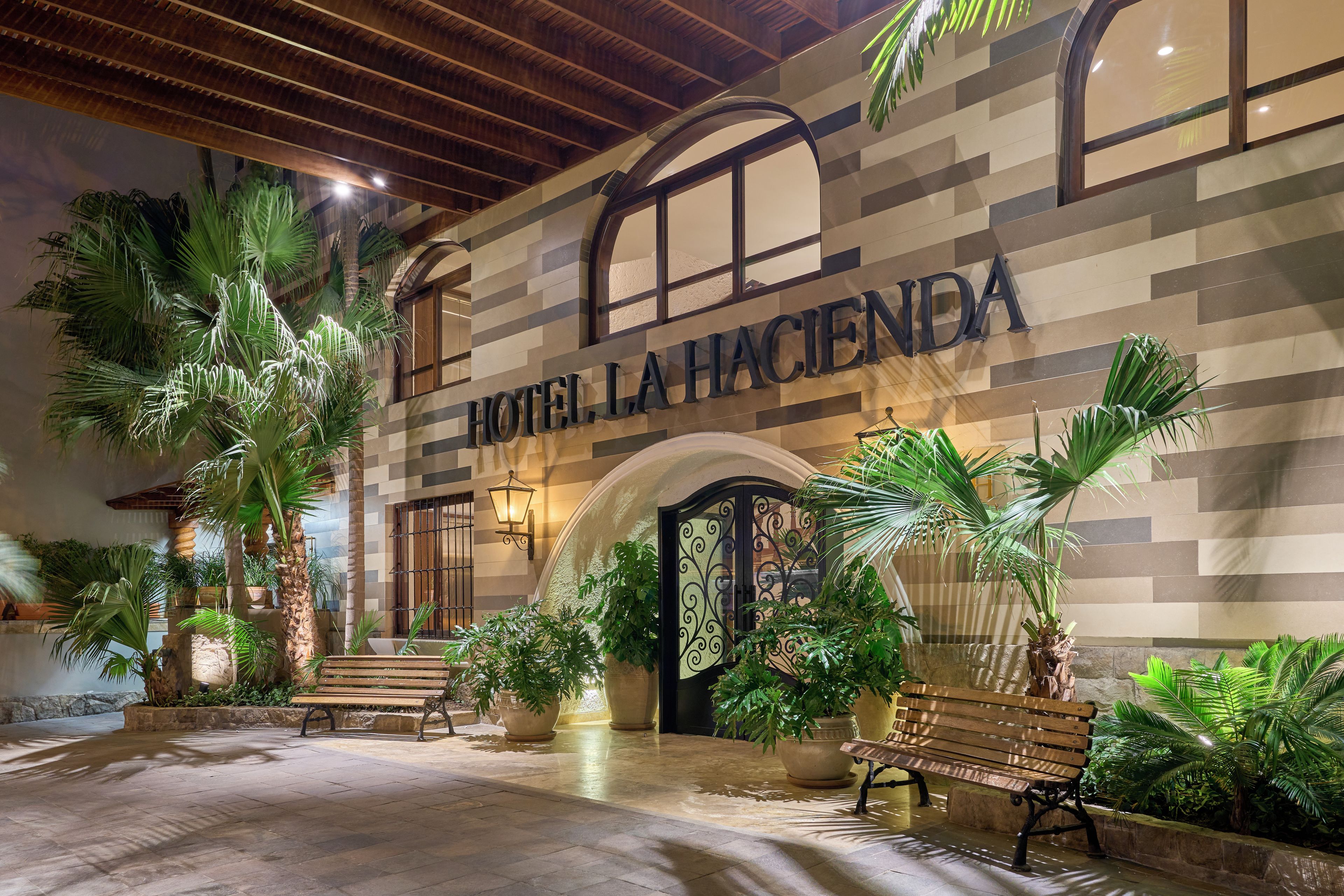 Entrada de un hotel con un letrero que dice 'HOTEL LA HACIENDA', rodeada de plantas en macetas y bancos.