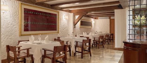 A dining area with wooden chairs and tables, a large abstract painting on the wall, and a view of greenery outside.