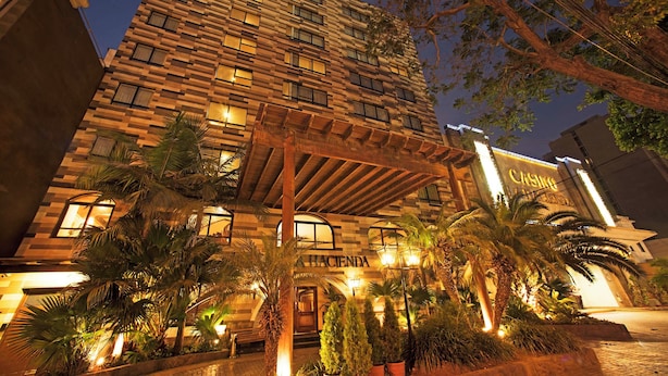 Um hotel com vários andares, fachada moderna, janelas iluminadas e área de entrada bem iluminada, com palmeiras e plantas em vasos.