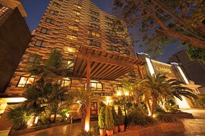 A multi-story hotel with a modern facade, illuminated windows, and a well-lit entrance area with palm trees and potted plants. - La Hacienda Miraflores (Lima)