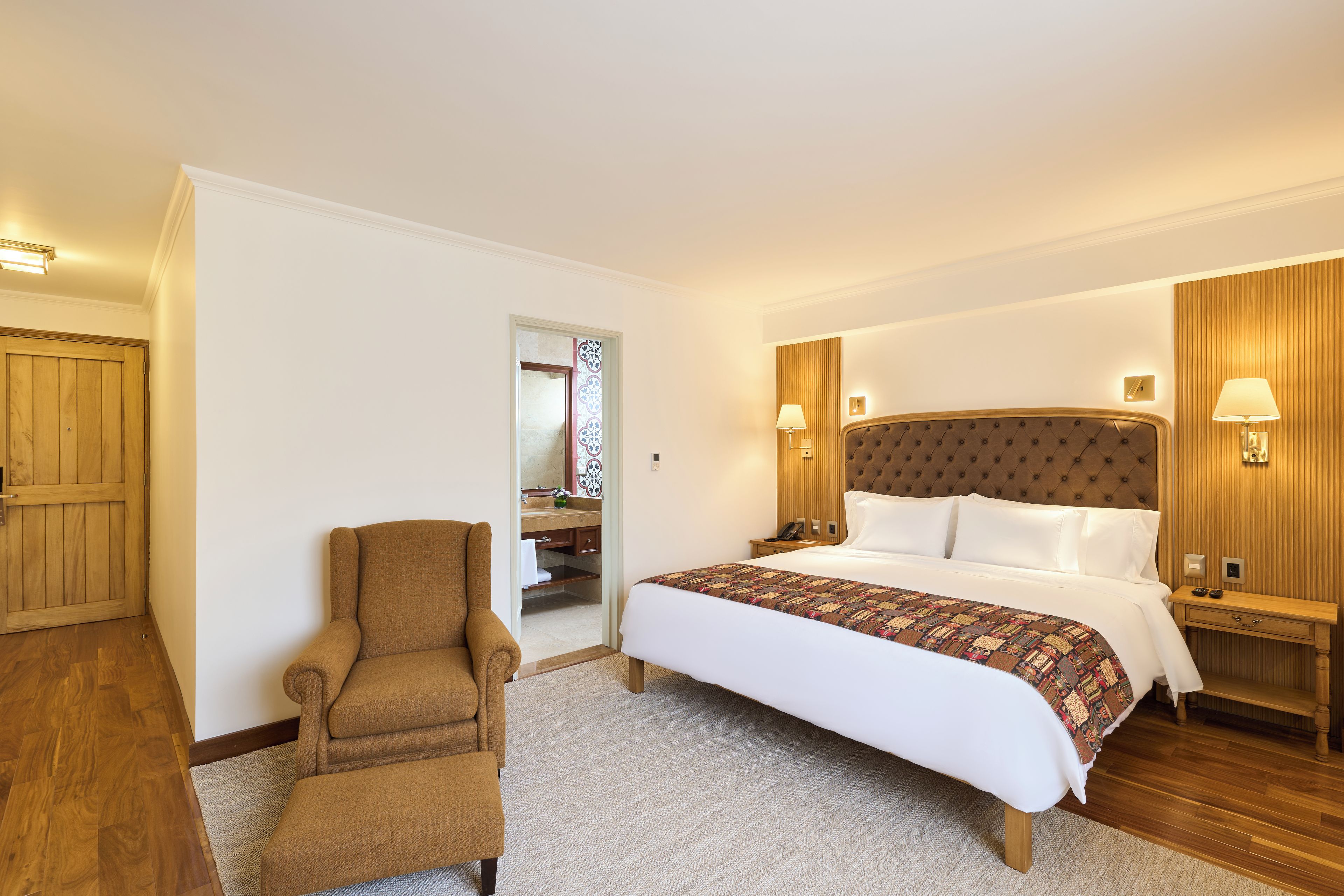 Una habitación de hotel con una cama grande, una zona de estar con una silla, un armario de madera y un baño visible a través de una puerta abierta.