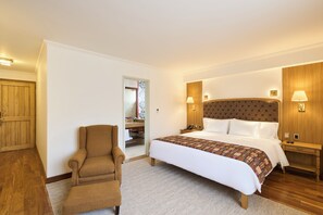 Une chambre d’hôtel avec un grand lit, un coin salon avec un fauteuil, une armoire en bois et une salle de bain visible à travers une porte ouverte.
