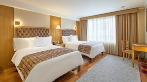 Quarto de hotel com duas camas, uma escrivaninha, uma cadeira e uma janela grande com cortinas.