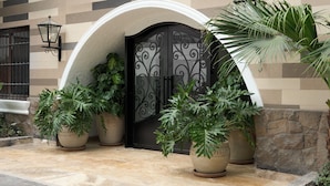 Entrada de um edifício com um portão preto ornamentado, plantas em vasos e piso de azulejos.