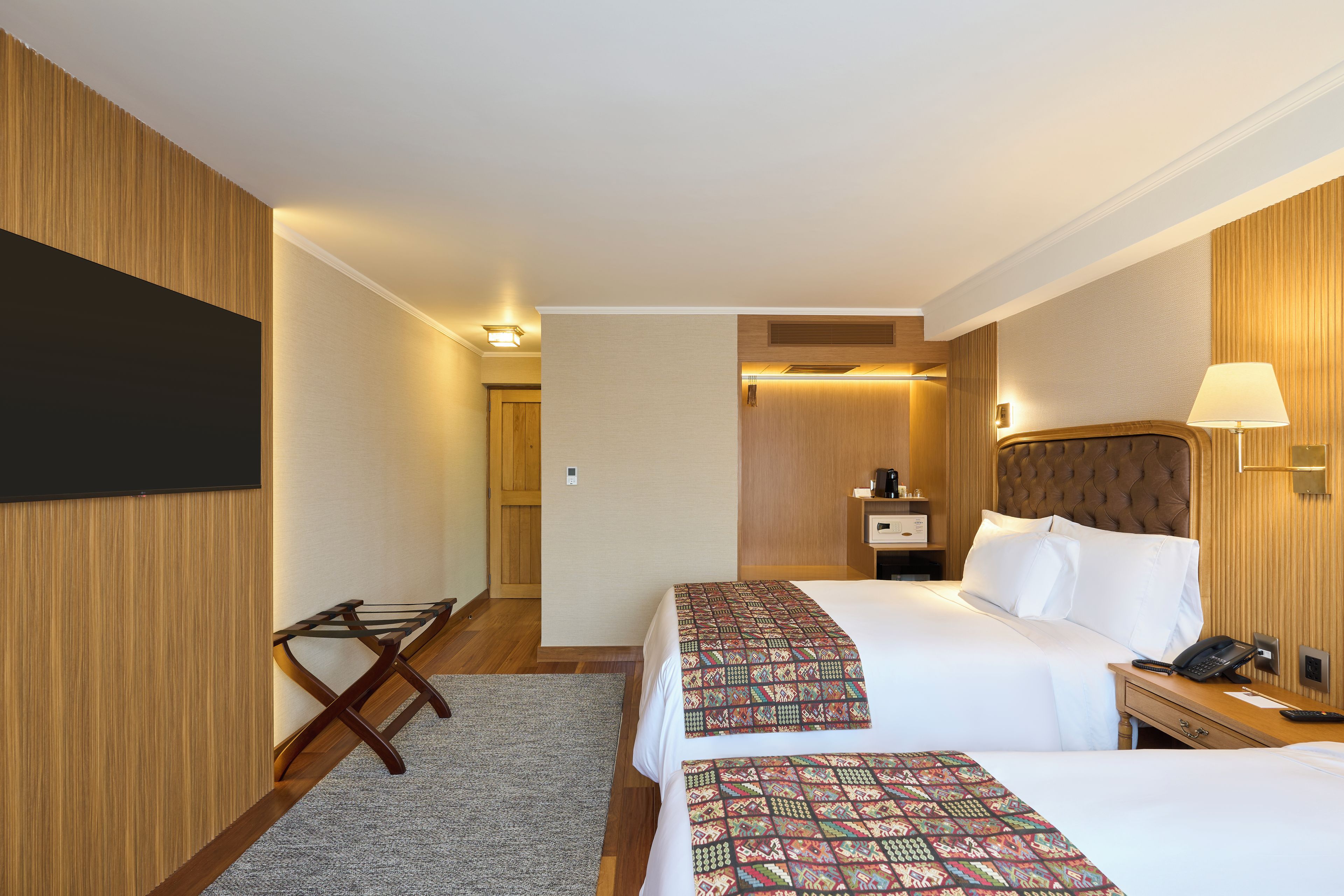 Habitación de hotel con dos camas, un escritorio, una silla, un televisor de pantalla plana y una mesita de noche con lámpara.