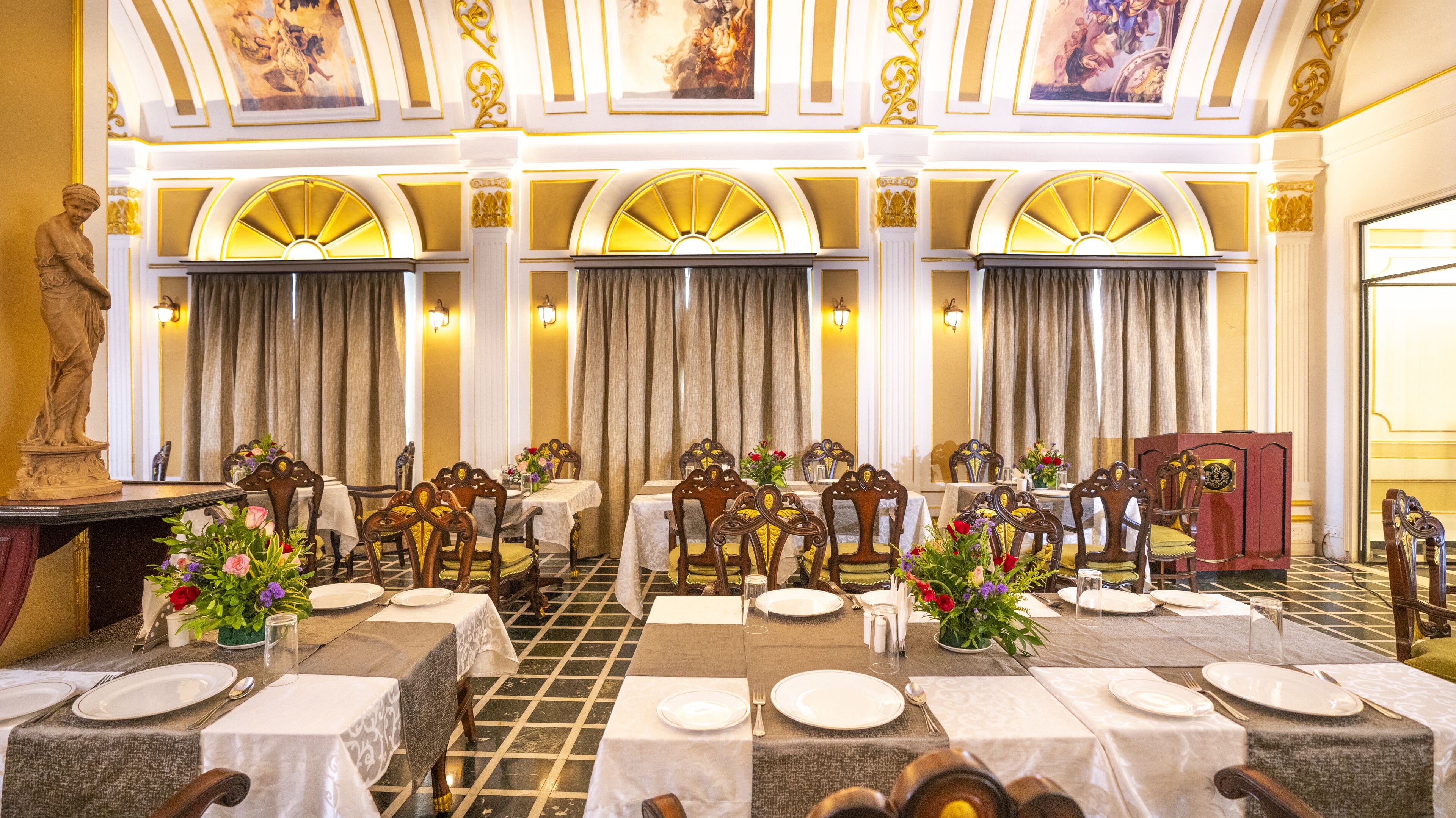 Una sala da pranzo formale con tavoli imbanditi, sedie decorate e opere d'arte classiche alle pareti.