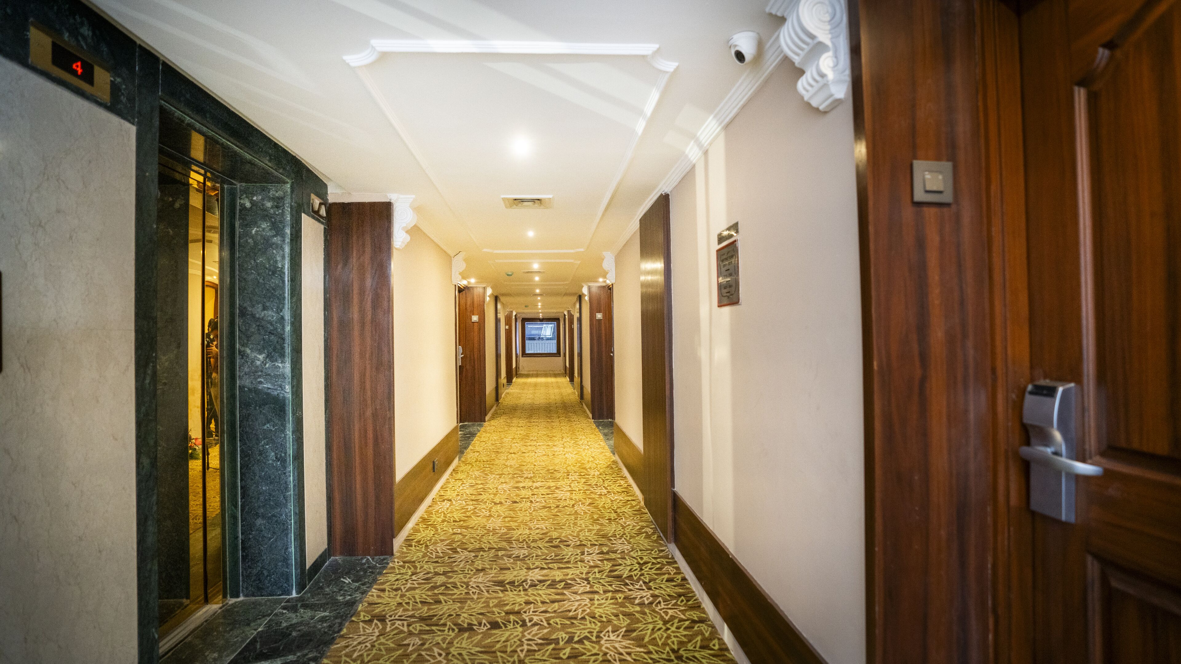 Un pasillo de hotel con alfombra estampada, puertas de madera y una cámara de seguridad.
