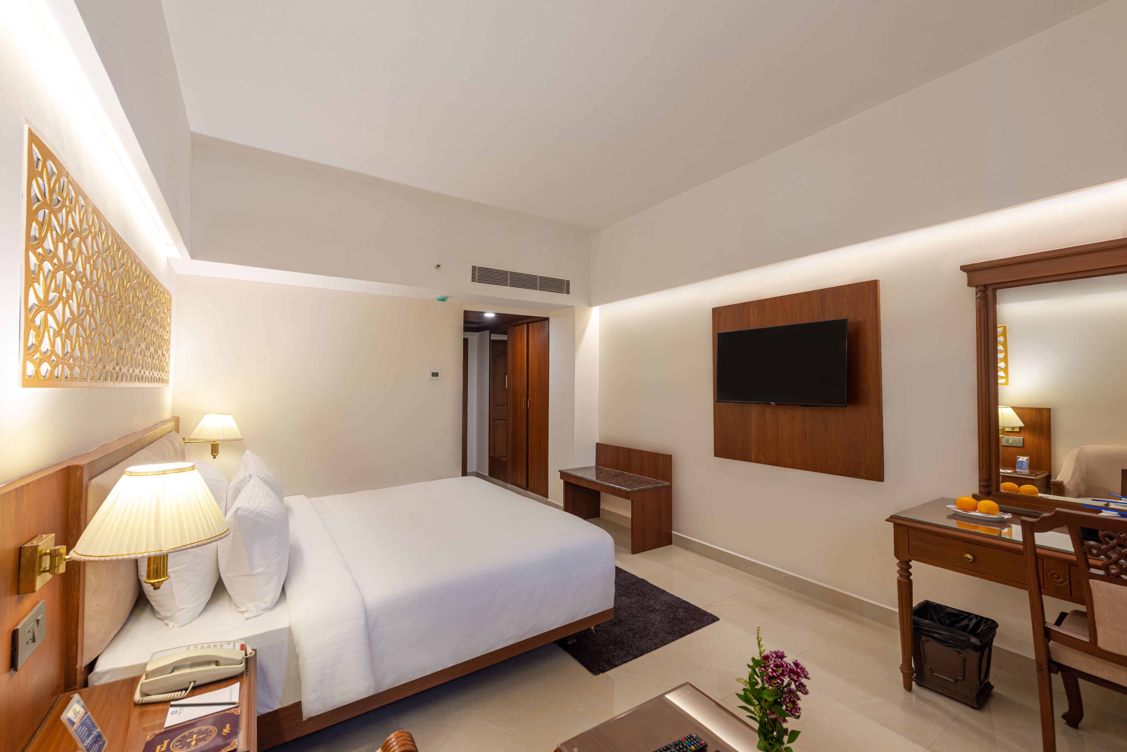 A hotel room with a bed, bedside table, lamp, TV, desk, chair, and a small trash bin.