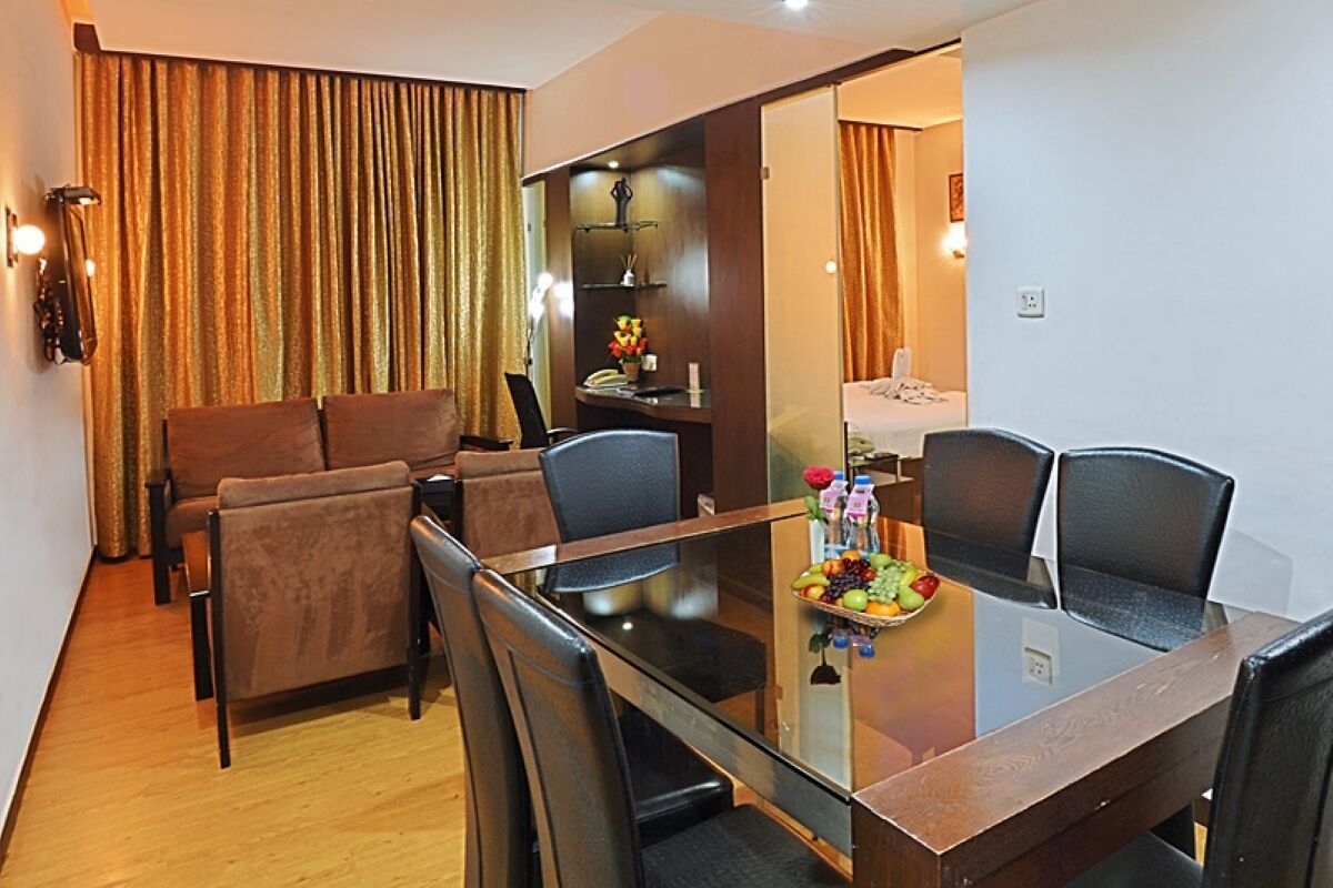 A hotel room with a glass dining table, a sofa, and a small kitchenette.