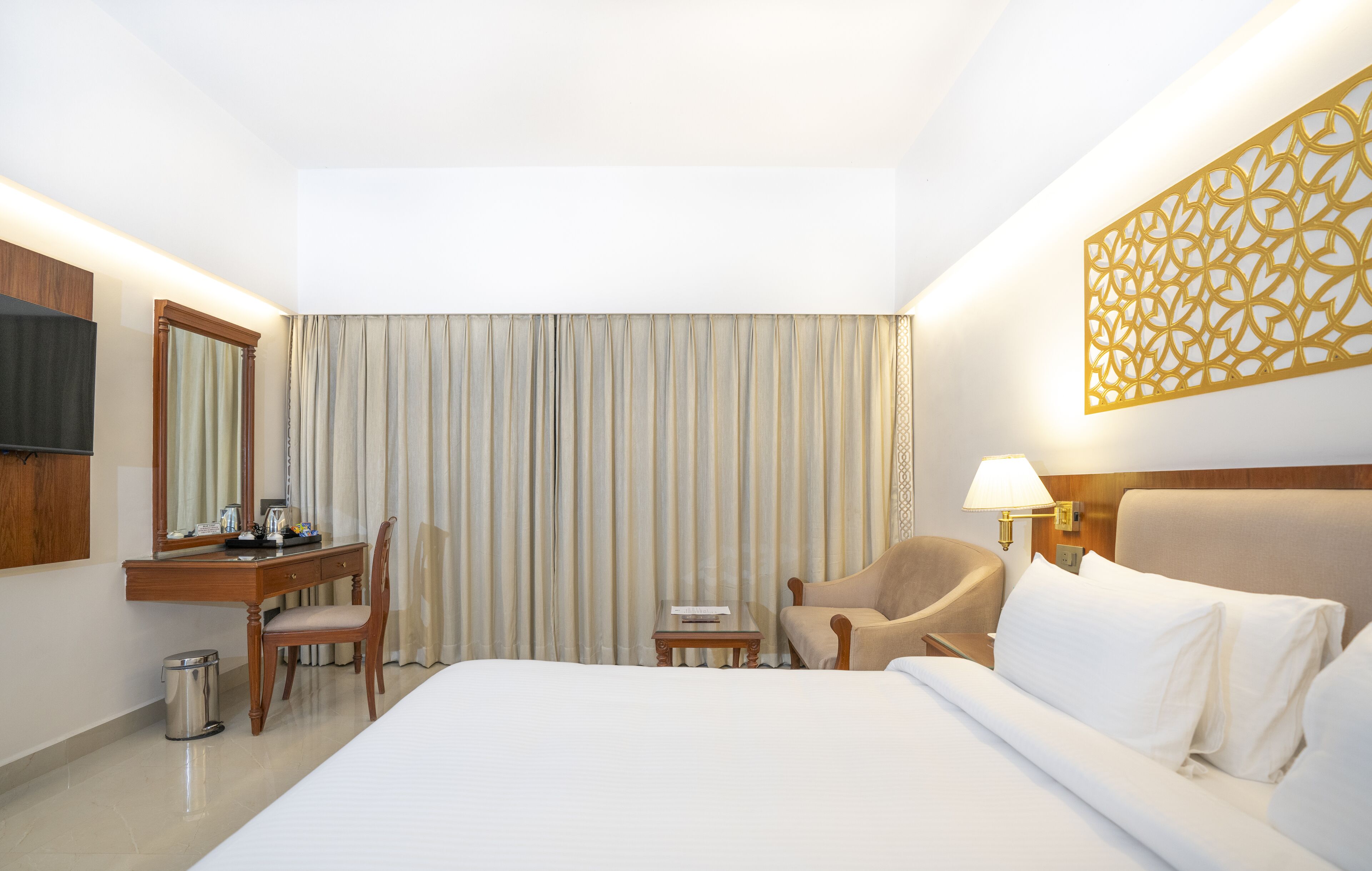 A hotel room with a bed, a desk with a chair, a small table, a lamp, and a decorative wall panel.