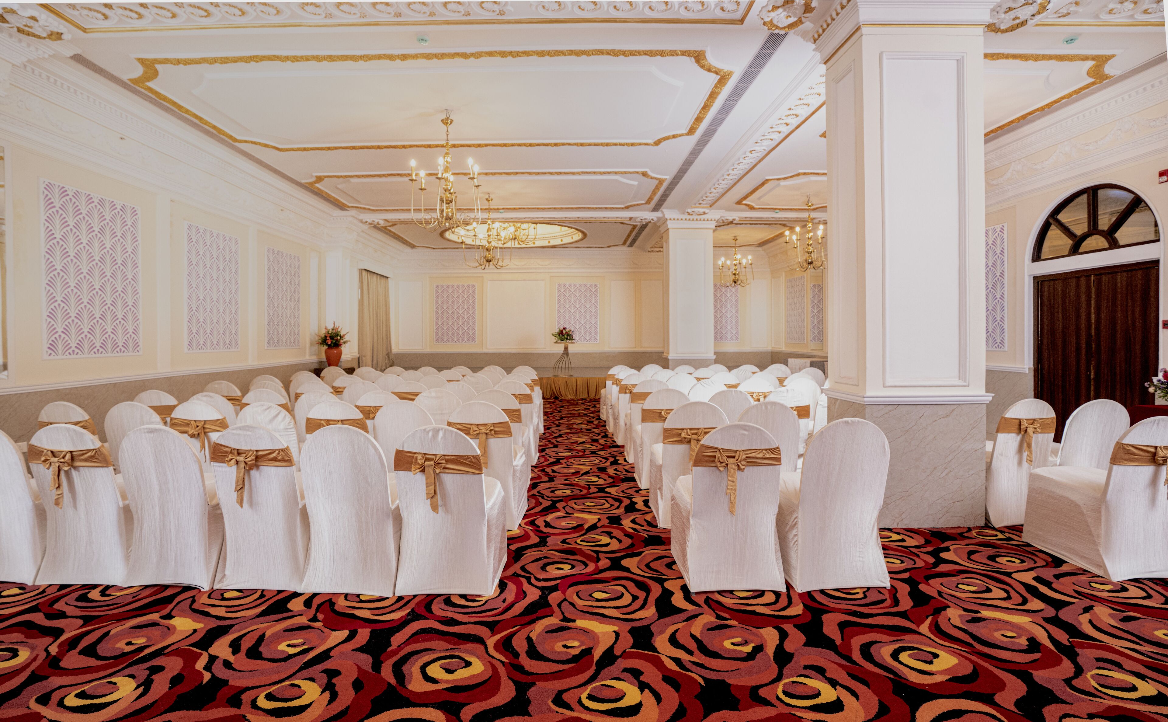 A banquet hall with rows of white chairs with gold sashes, a red and black patterned carpet, and ornate ceiling decorations.