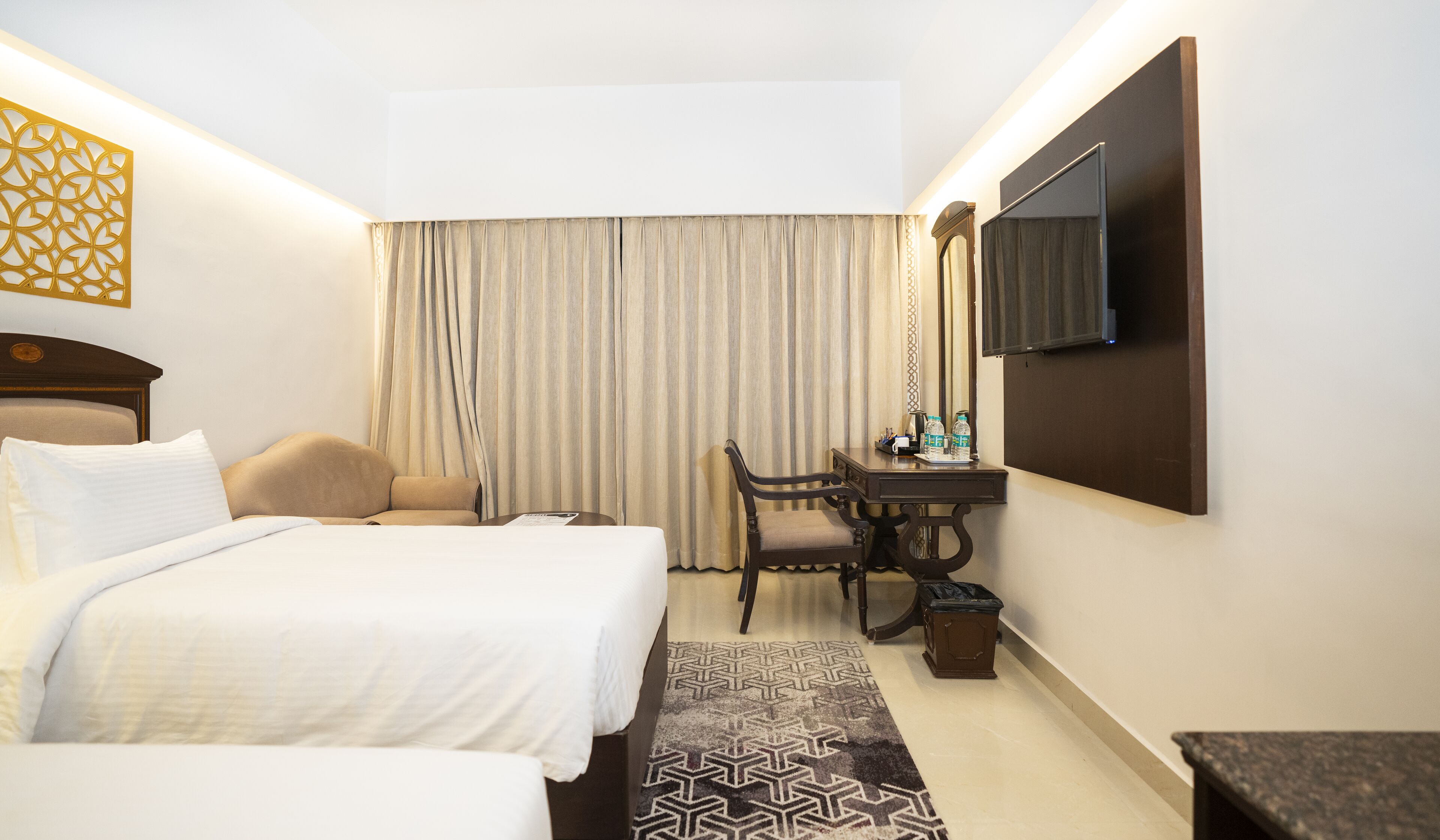 A hotel room with a bed, a desk, a chair, a TV, and a patterned rug.