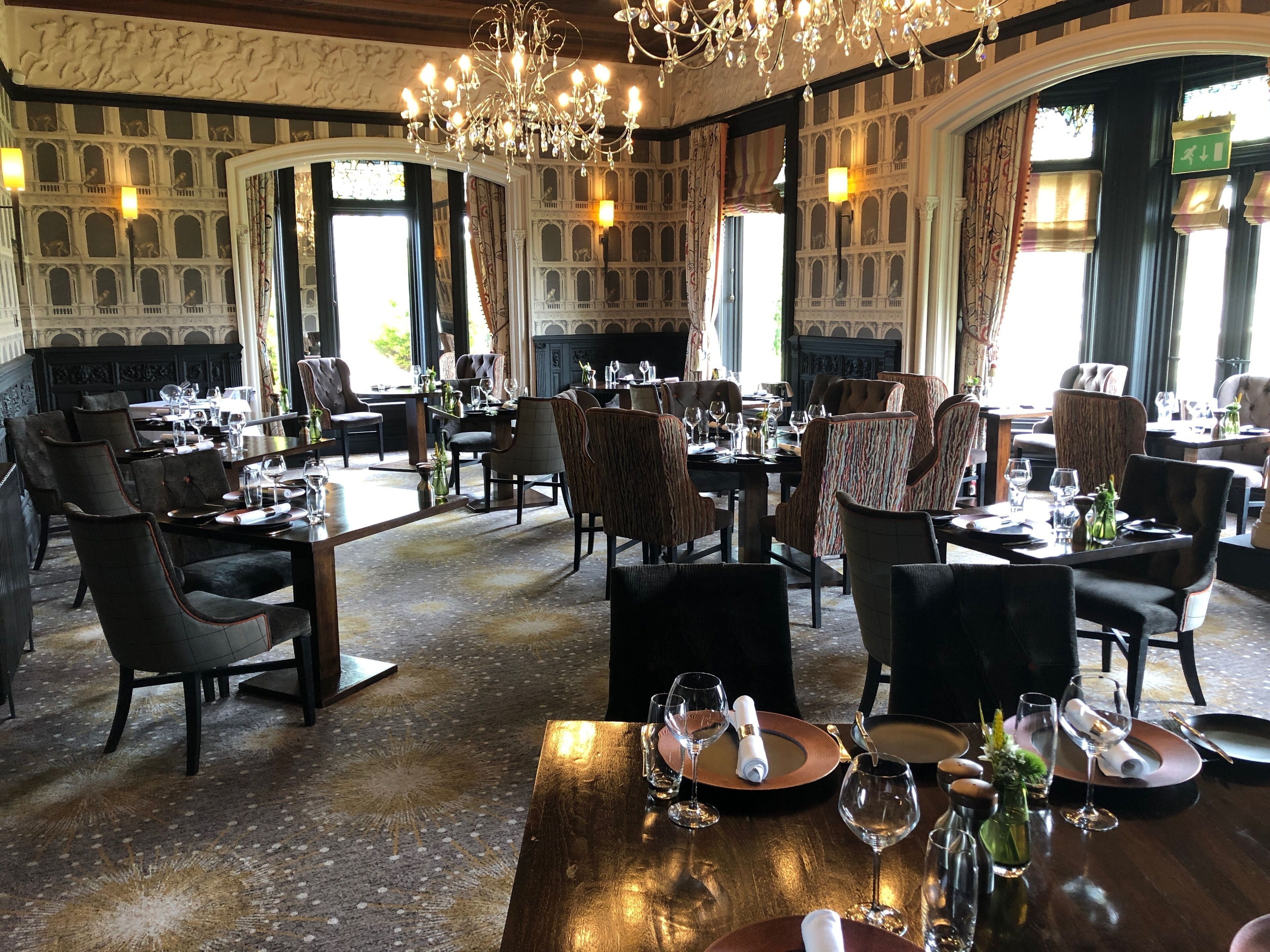 A restaurant with multiple dining tables and chairs, large windows with curtains, and a chandelier.
