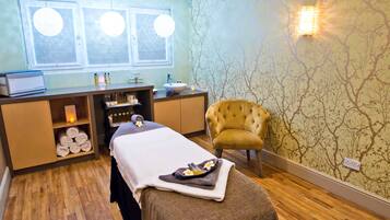 A spa room with a massage table, a chair, a vanity with toiletries, and a window with blinds.