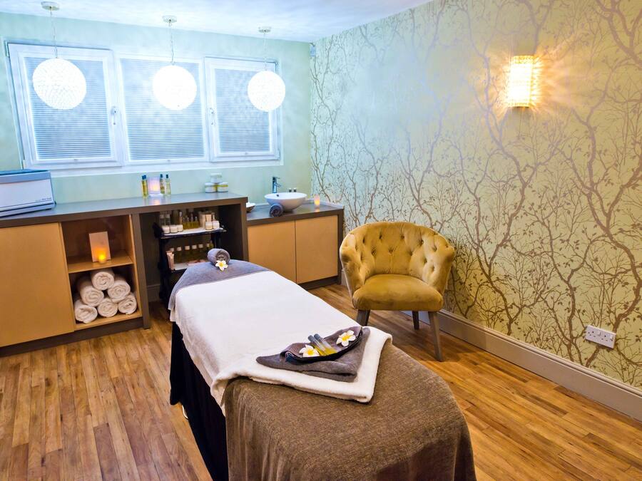 A spa room with a massage table, a chair, a vanity with toiletries, and a window with blinds.