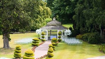A gazebo with a tiled roof surrounded by neatly trimmed hedges and a row of chairs.