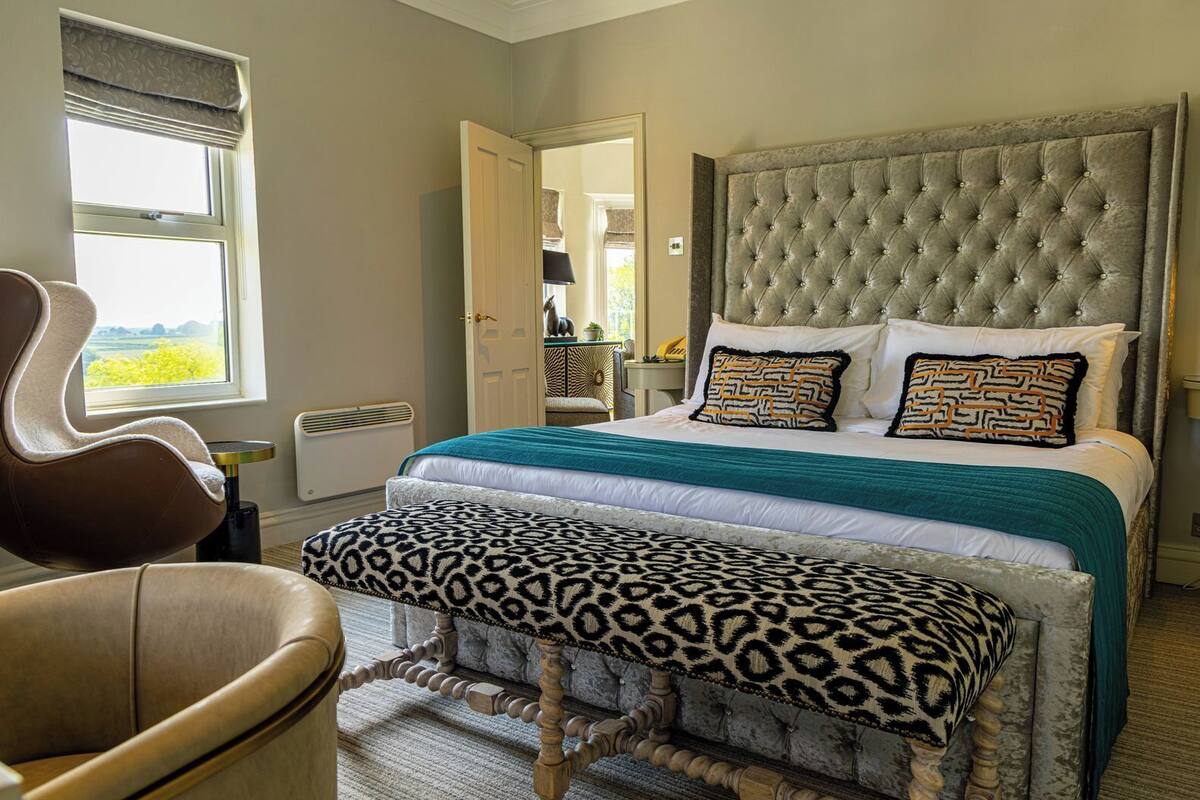 A bedroom with a tufted headboard, a bed with patterned pillows, a leopard print bench, and a chair by a window.