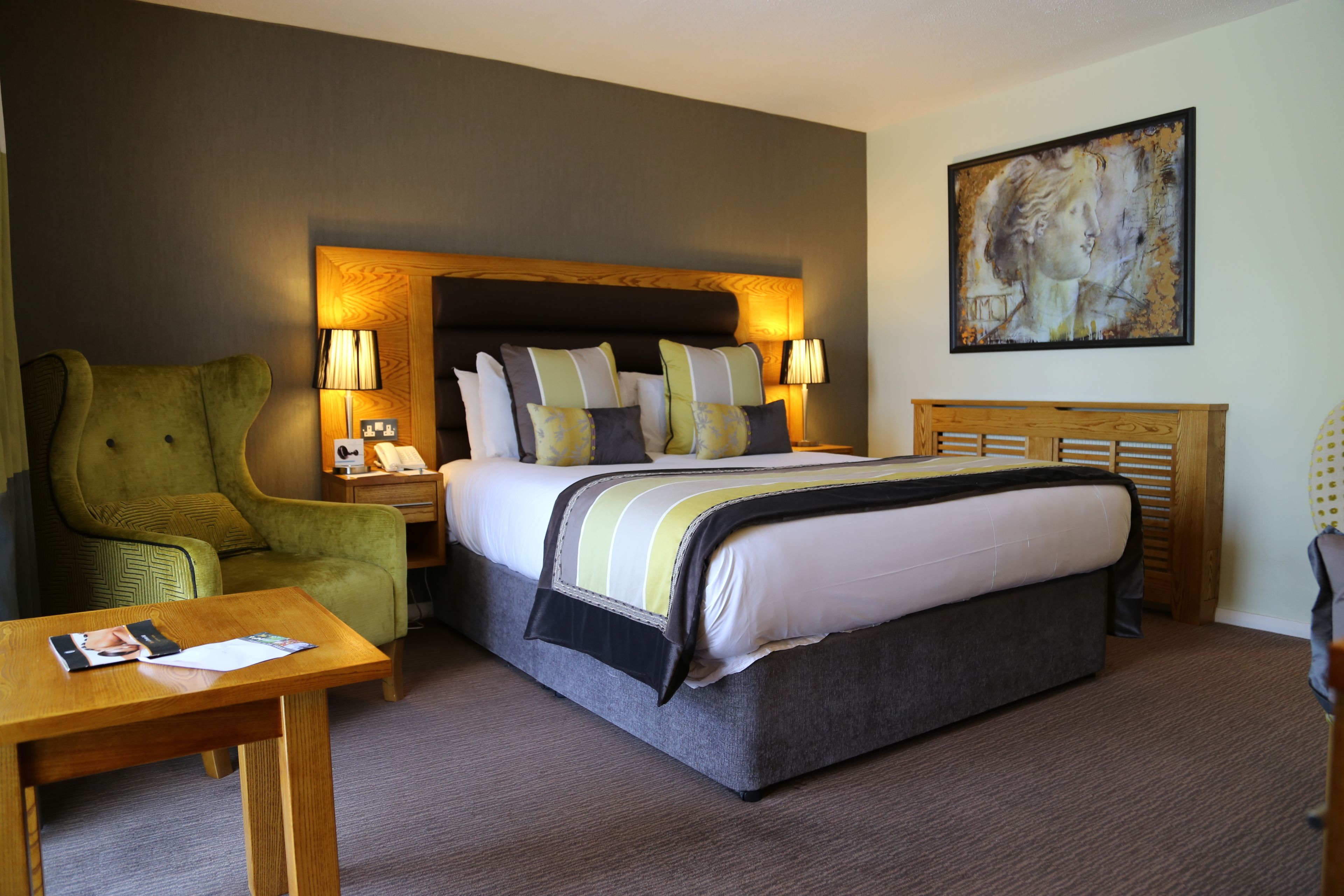 A hotel room with a large bed, a green armchair, a wooden nightstand, and a painting on the wall.