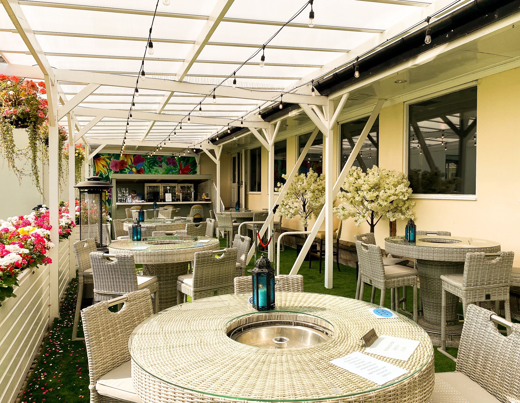 Een terras buiten met rieten meubilair, een ronde tafel in het midden en hangmanden met bloemen.