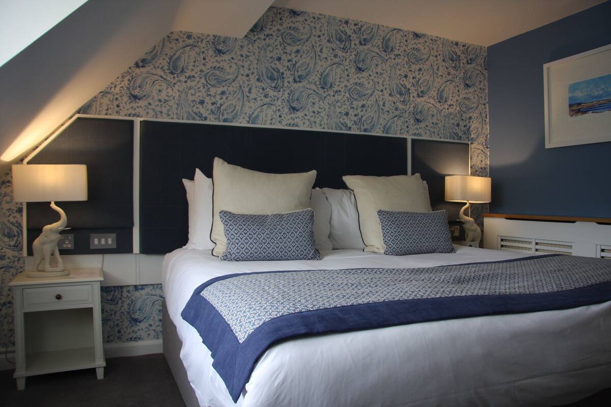 A bedroom with a bed, two bedside tables, a headboard, and a wall with a patterned wallpaper.