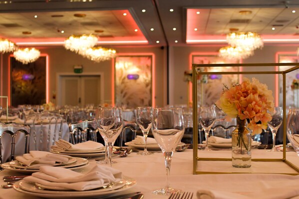 Une salle de banquet où les tables sont dressées pour un événement formel, avec de la verrerie, des serviettes pliées et un vase rempli de fleurs.