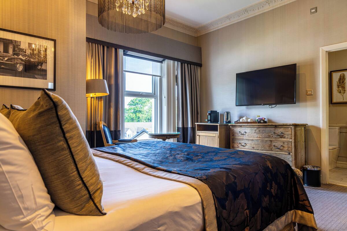 A hotel room with a large bed, a television mounted on the wall, a desk with a lamp, and a window with curtains.