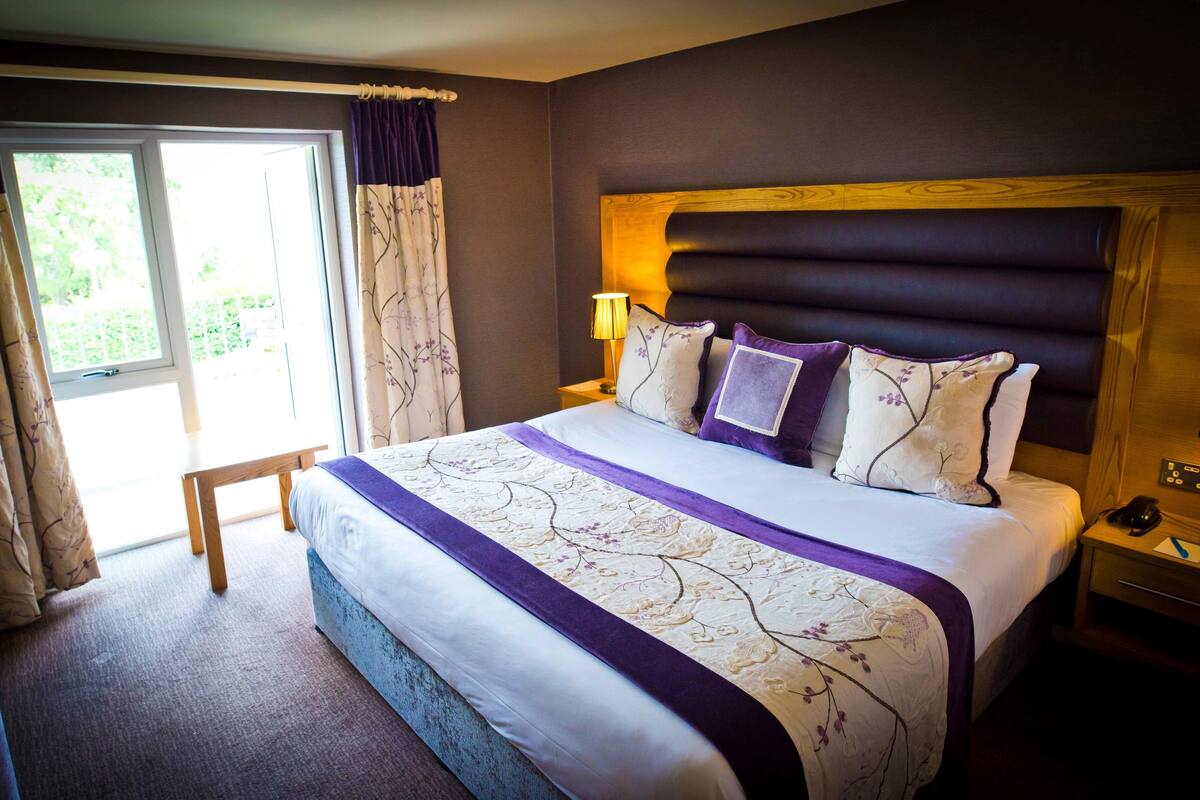 A neatly made bed with a headboard, a bedside table with a lamp, and a window with curtains.