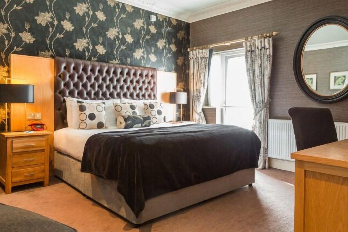 A bedroom with a tufted headboard, a bed with patterned pillows, a bedside table with a lamp, a chair, and a large mirror on the wall.