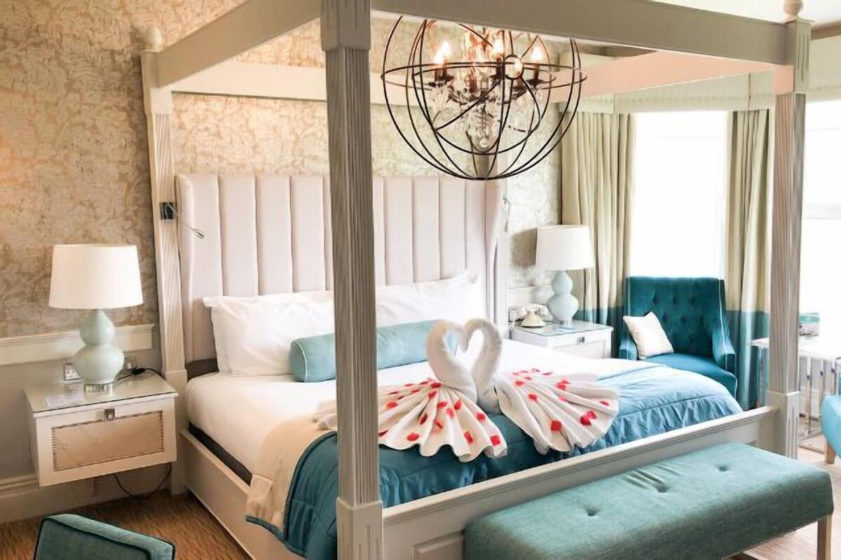 A four-poster bed with a chandelier, a blue upholstered bench, and a heart-shaped decoration on the bed.