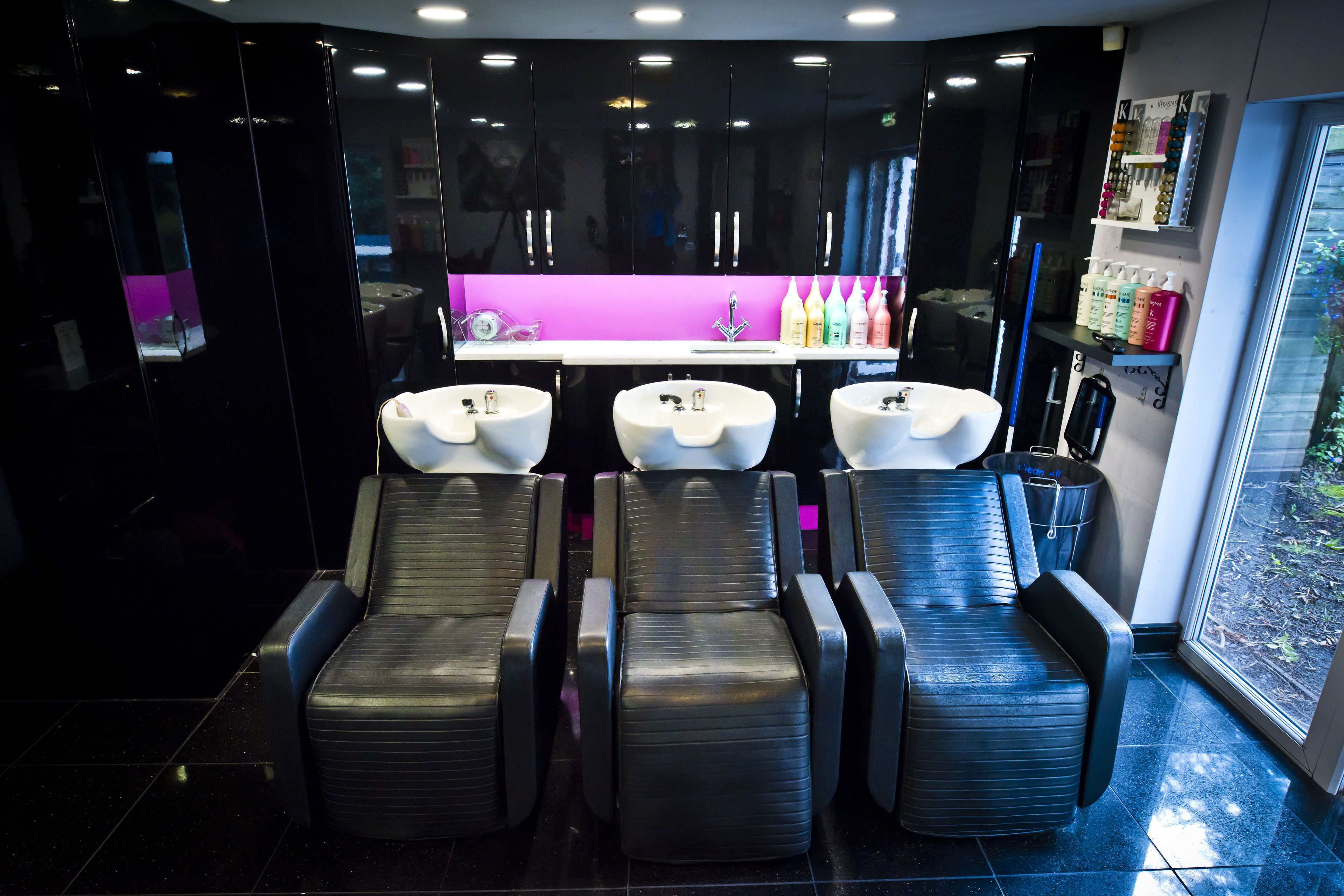 Ein moderner Salon mit drei schwarzen Massagesesseln, einer zentralen Theke mit beleuchteter Violett-Präsentation und einem Fenster mit Blick auf Grünflächen.