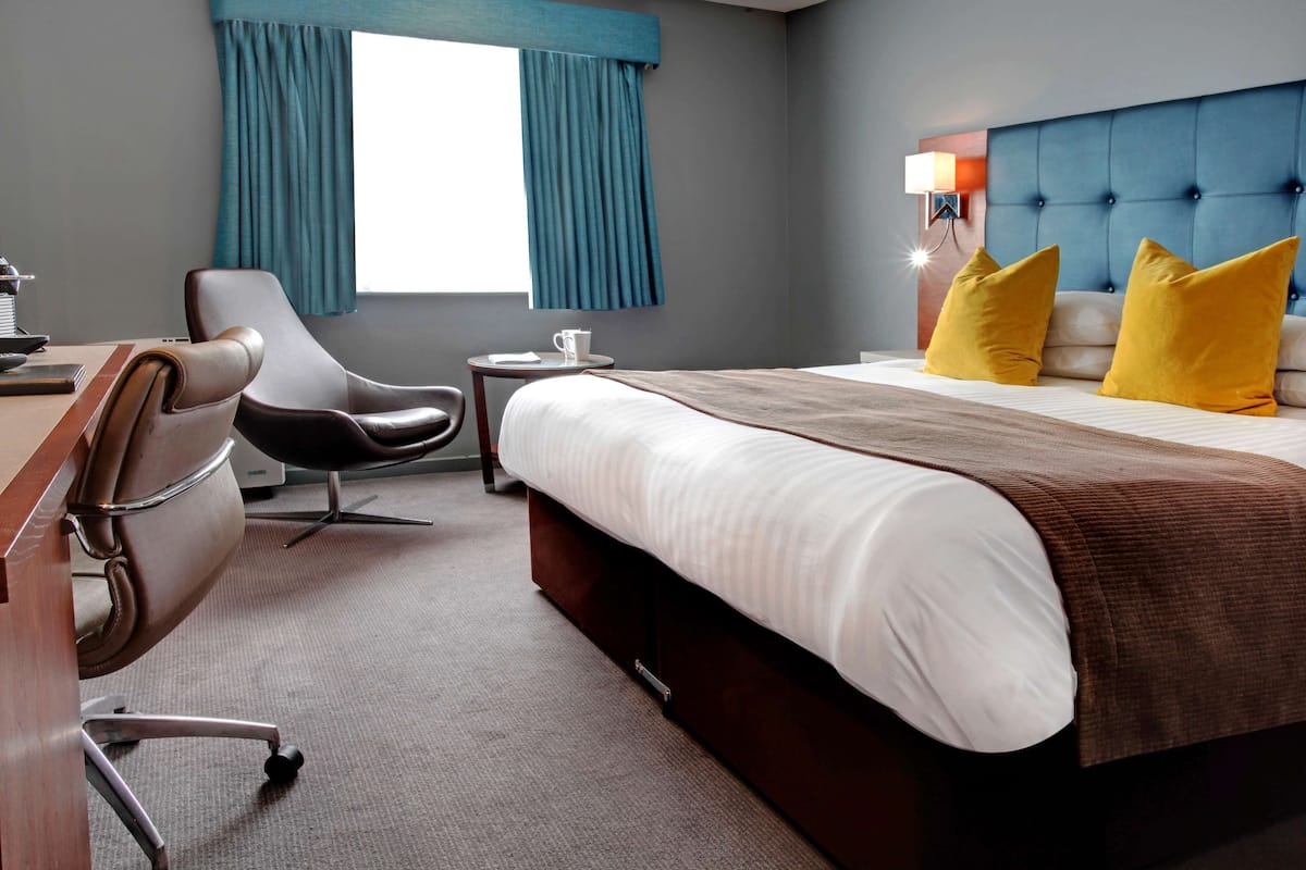 A hotel room with a bed, a desk, a chair, a small table, and a window with curtains.