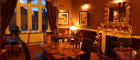 Un comedor cálidamente iluminado con chimenea, ventanas grandes con cortinas y cuadros en las paredes.